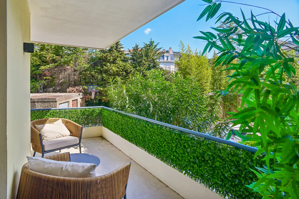 Balcony with two wicker chairs, a small table, and a green hedge on the railing, overlooking lush trees.