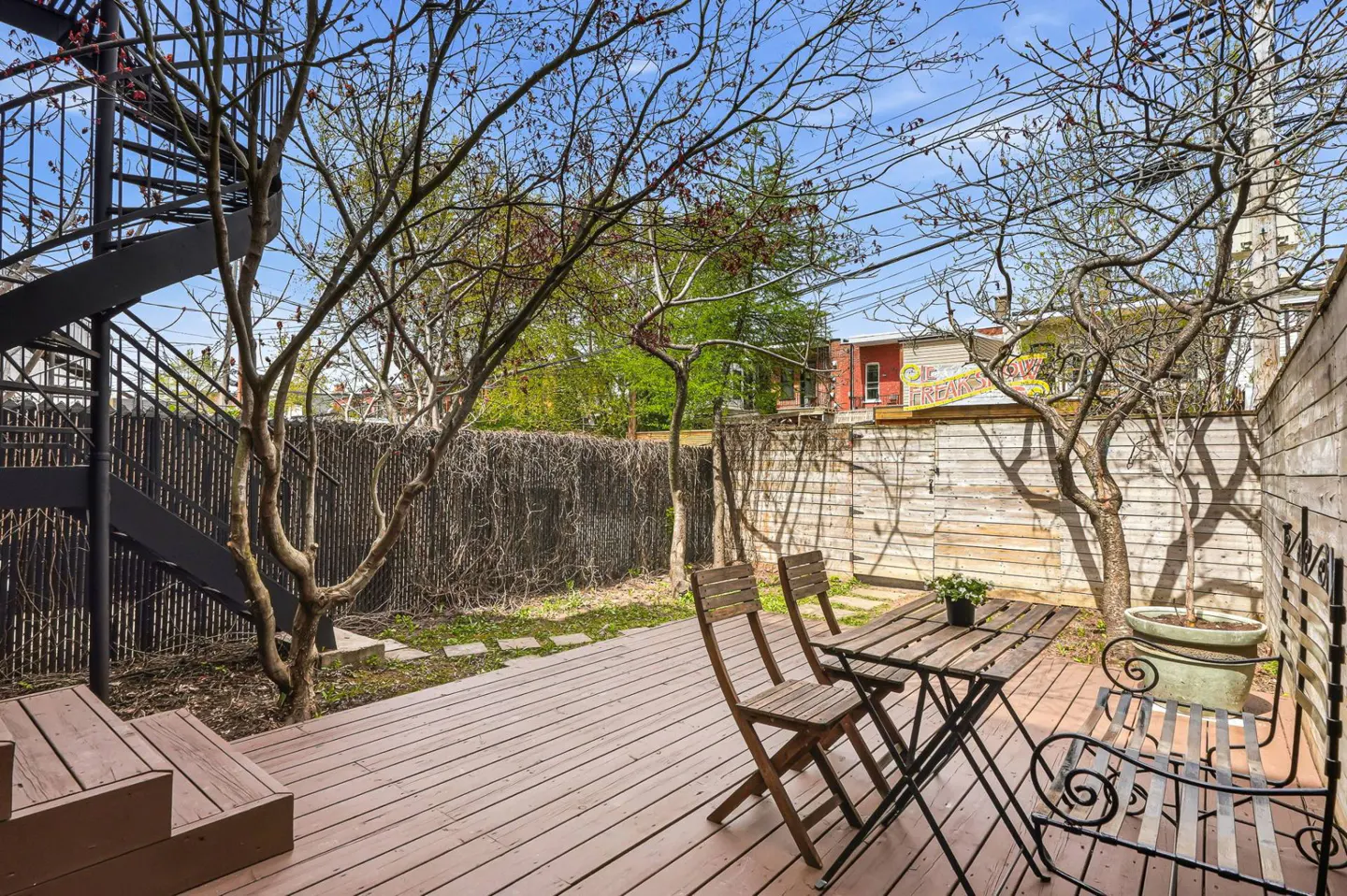 A backyard patio with a wooden deck, table, chairs, and a black spiral staircase. Trees and a fence surround the space.
