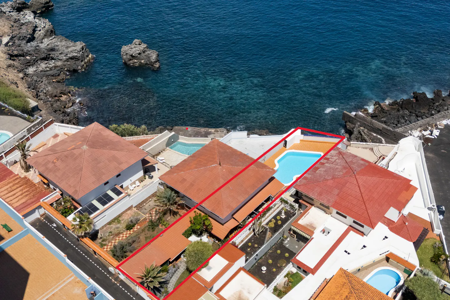 Aerial view of a coastal property with red-tiled roofs, a blue pool, and a red boundary line, set against a backdrop of the ocean and rocky cliffs.