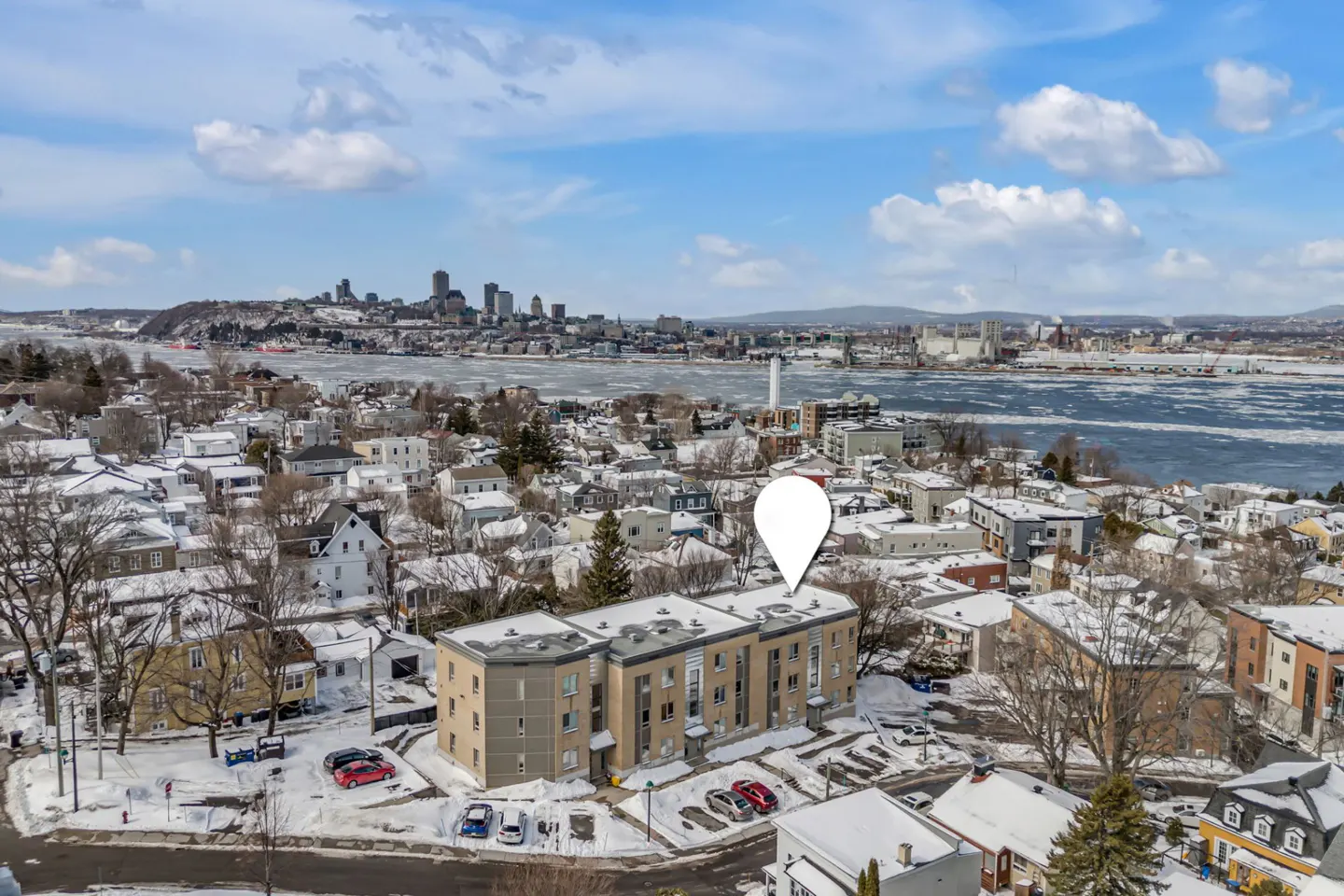 Aerial view of a beige apartment building with a white location pin, surrounded by snow-covered houses and a distant city skyline.