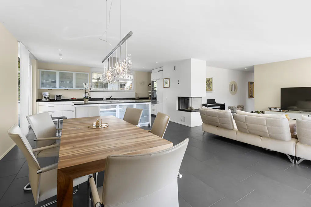 Open-concept living space with a wood dining table, beige chairs, and a crystal chandelier. White kitchen, fireplace, and sofa in the background.
