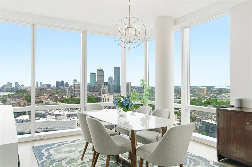 Bright dining room with city view. White table with six gray chairs, globe chandelier, and blue floral centerpiece. Large windows overlook Boston skyline.