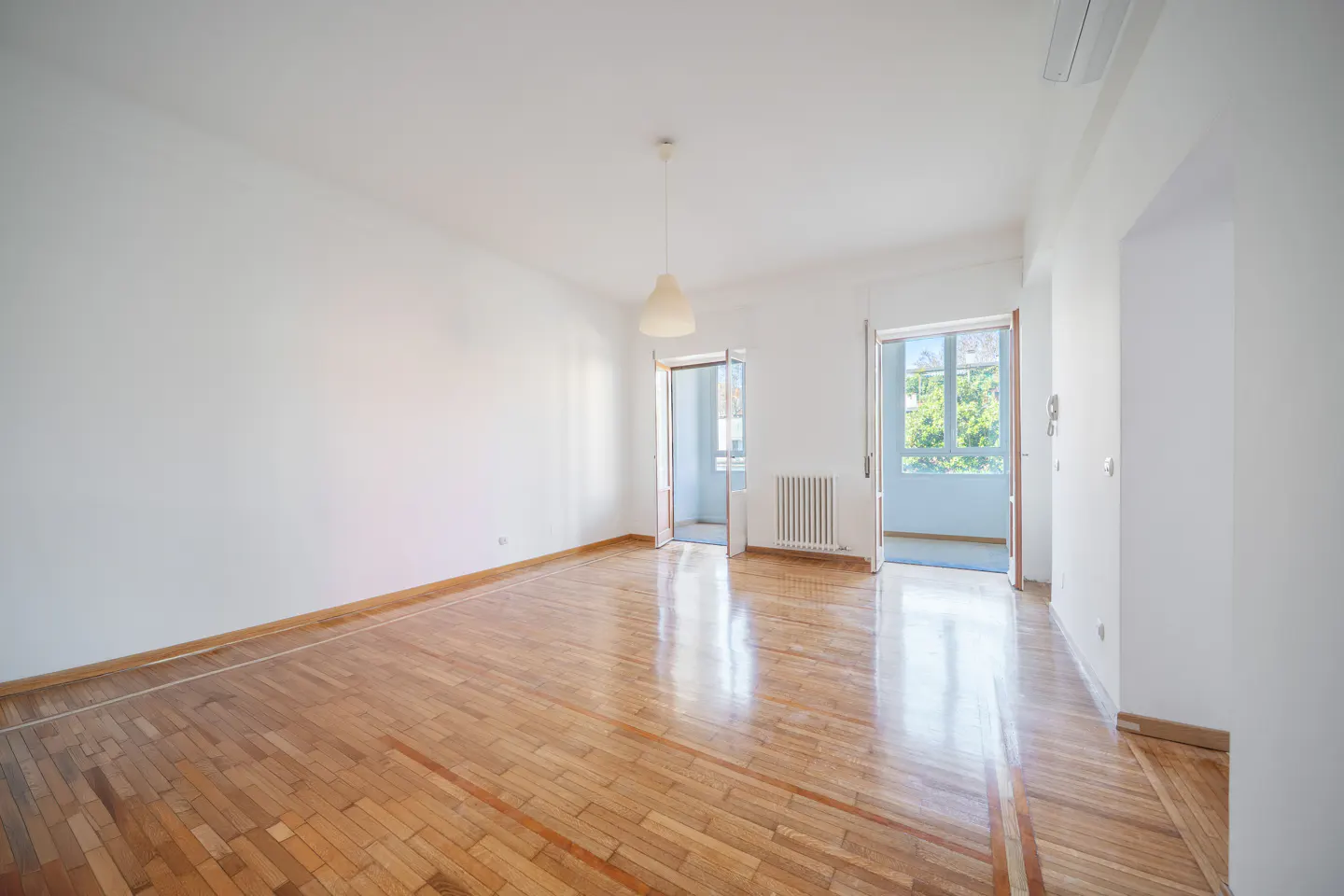 Bright, empty room with white walls, wood floors, and open doors to a balcony with green trees visible.