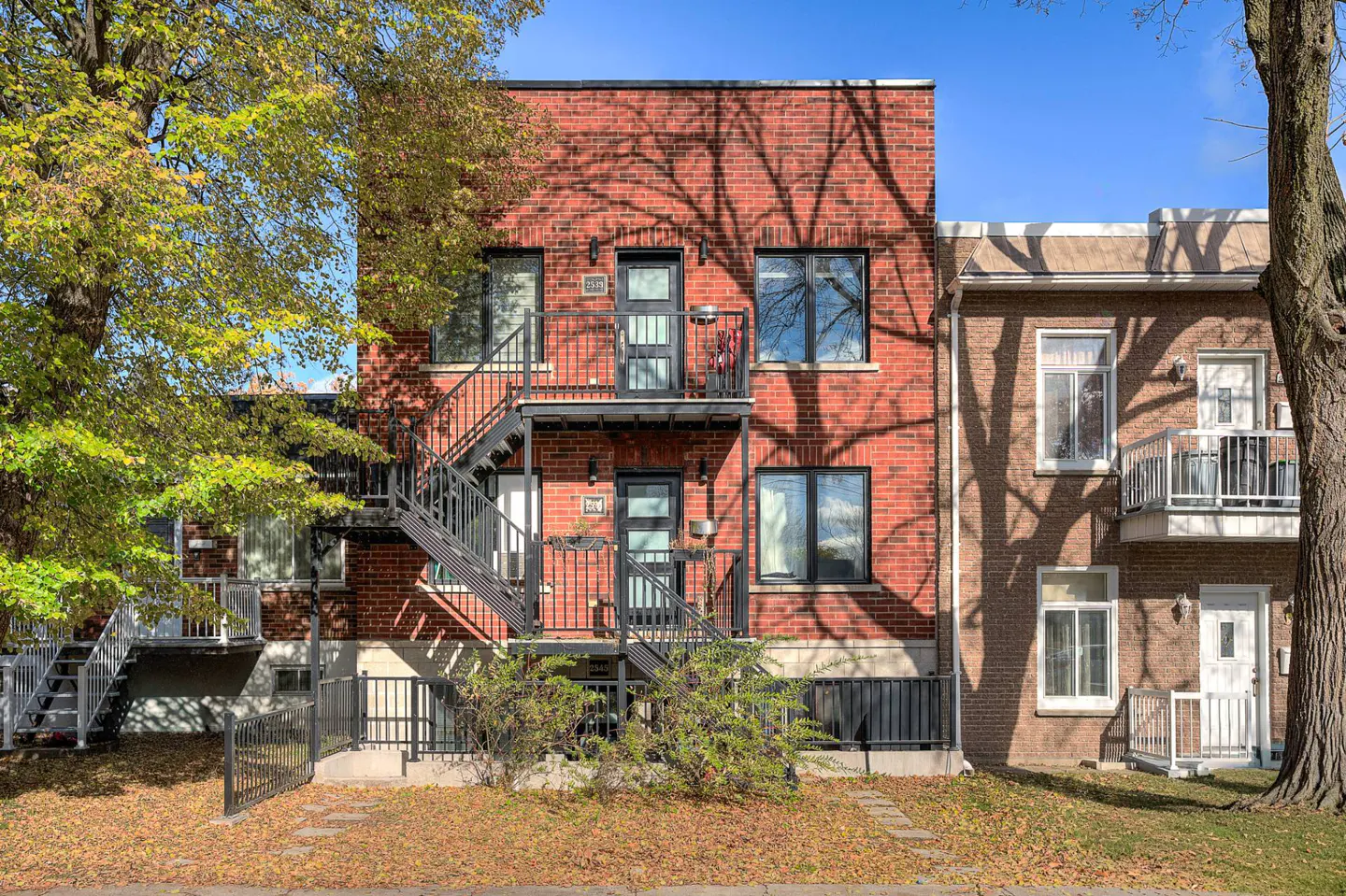 Red brick apartment building with black metal stairs and balconies. Trees with yellow leaves frame the building.