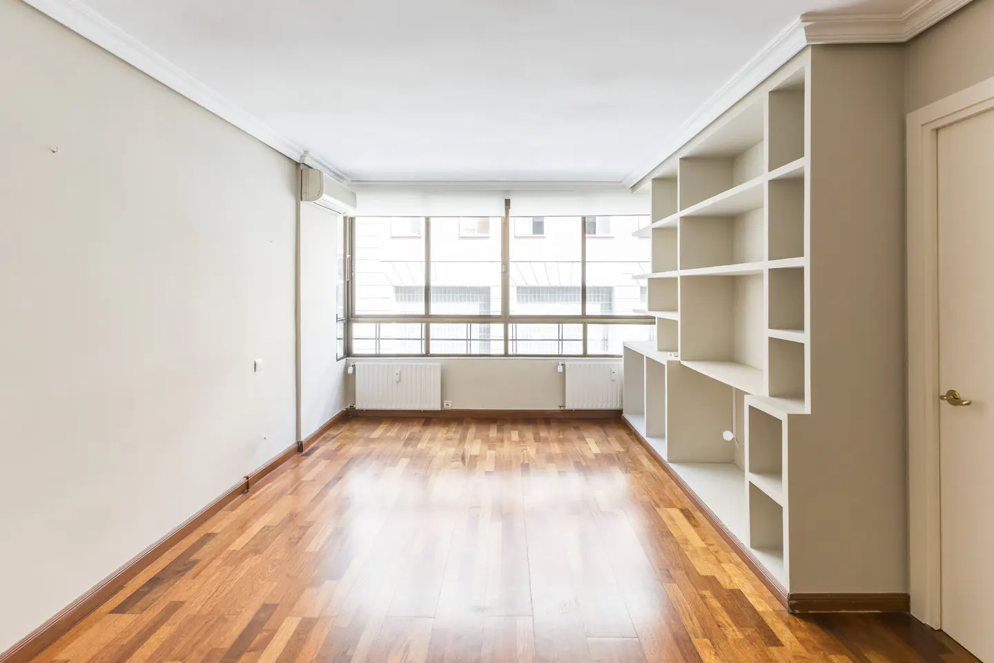 Bright, empty room with hardwood floors, a large window, and a built-in white bookshelf.