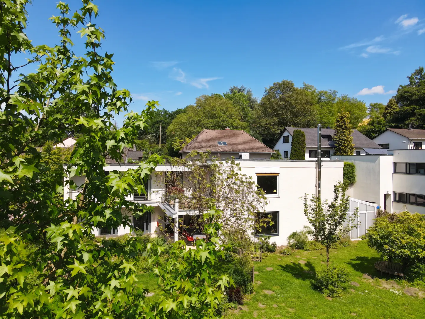 Two-story white house with a balcony, surrounded by green trees and a lawn under a blue sky.