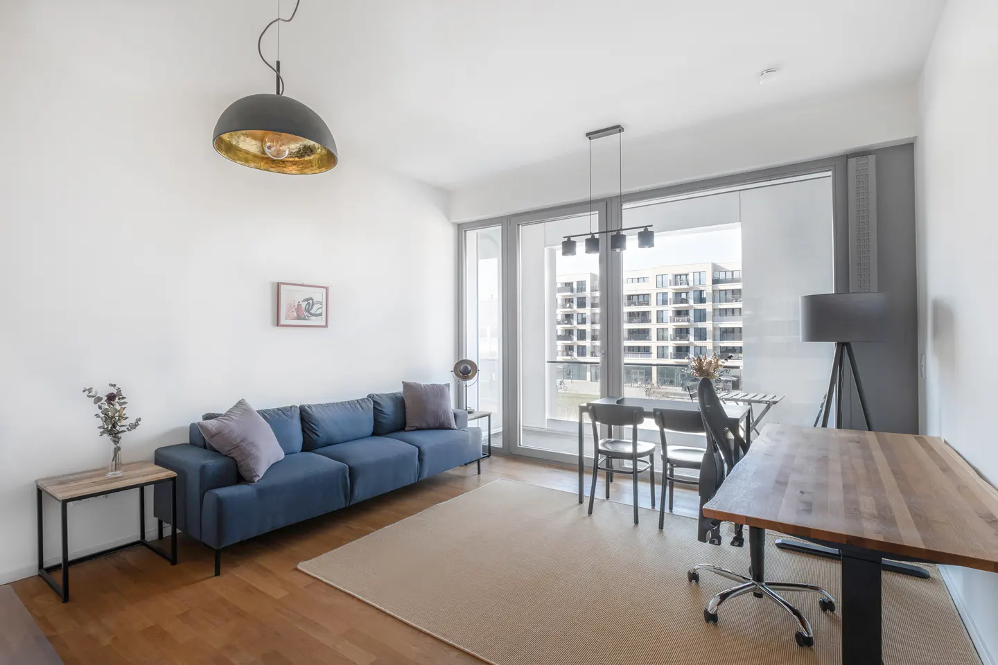 Bright living room with a blue sofa, wooden floors, and a large window overlooking a city building. A dining table and desk are also present.