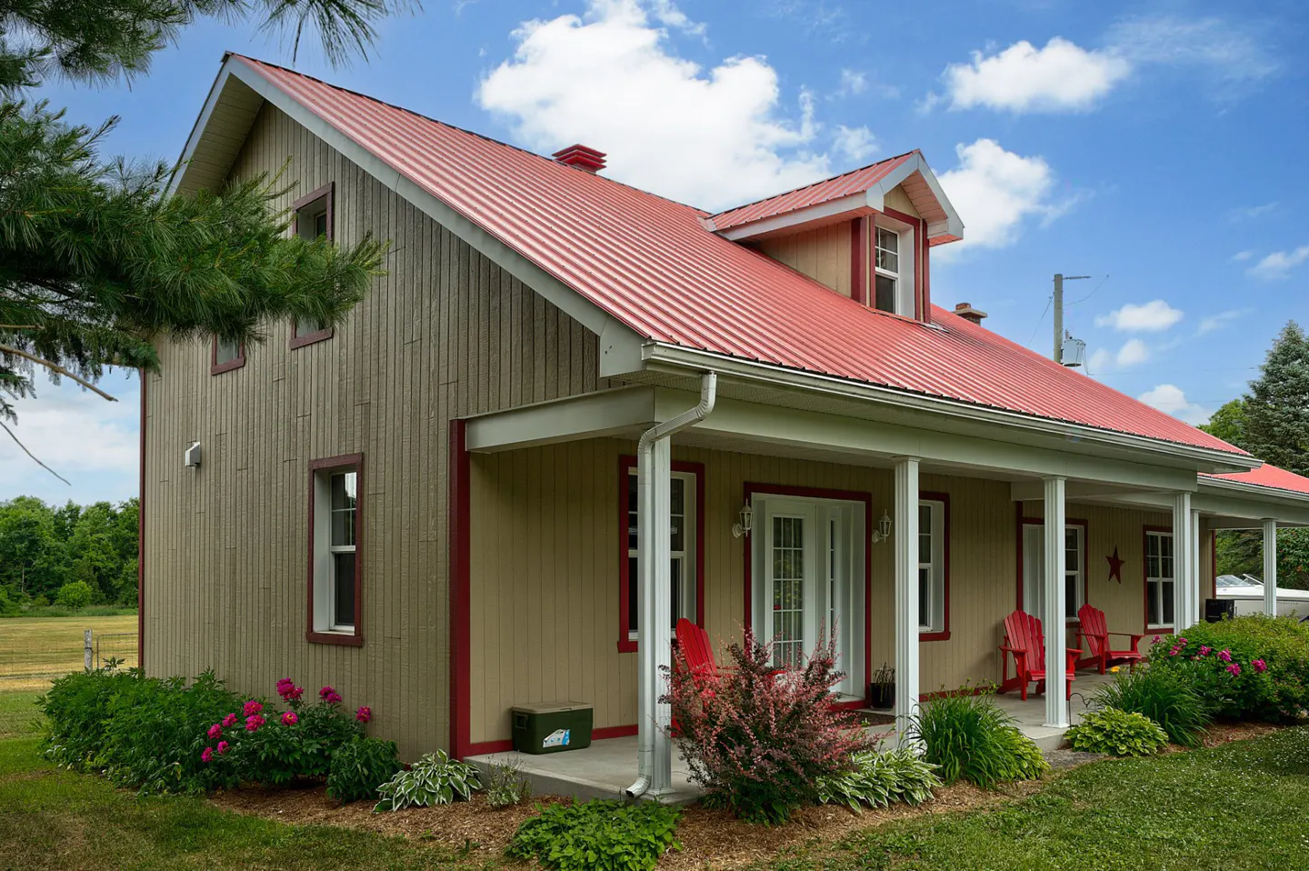Tan house with a red metal roof and a white porch with red chairs, surrounded by green grass and plants.