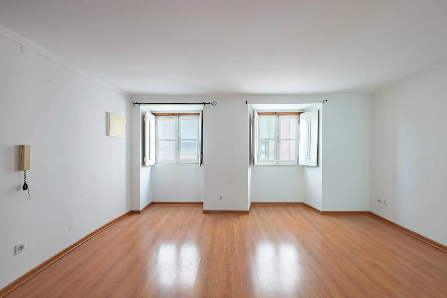 Bright, empty room with wood floors, white walls, and two open windows with white frames. A wall-mounted phone is on the left.
