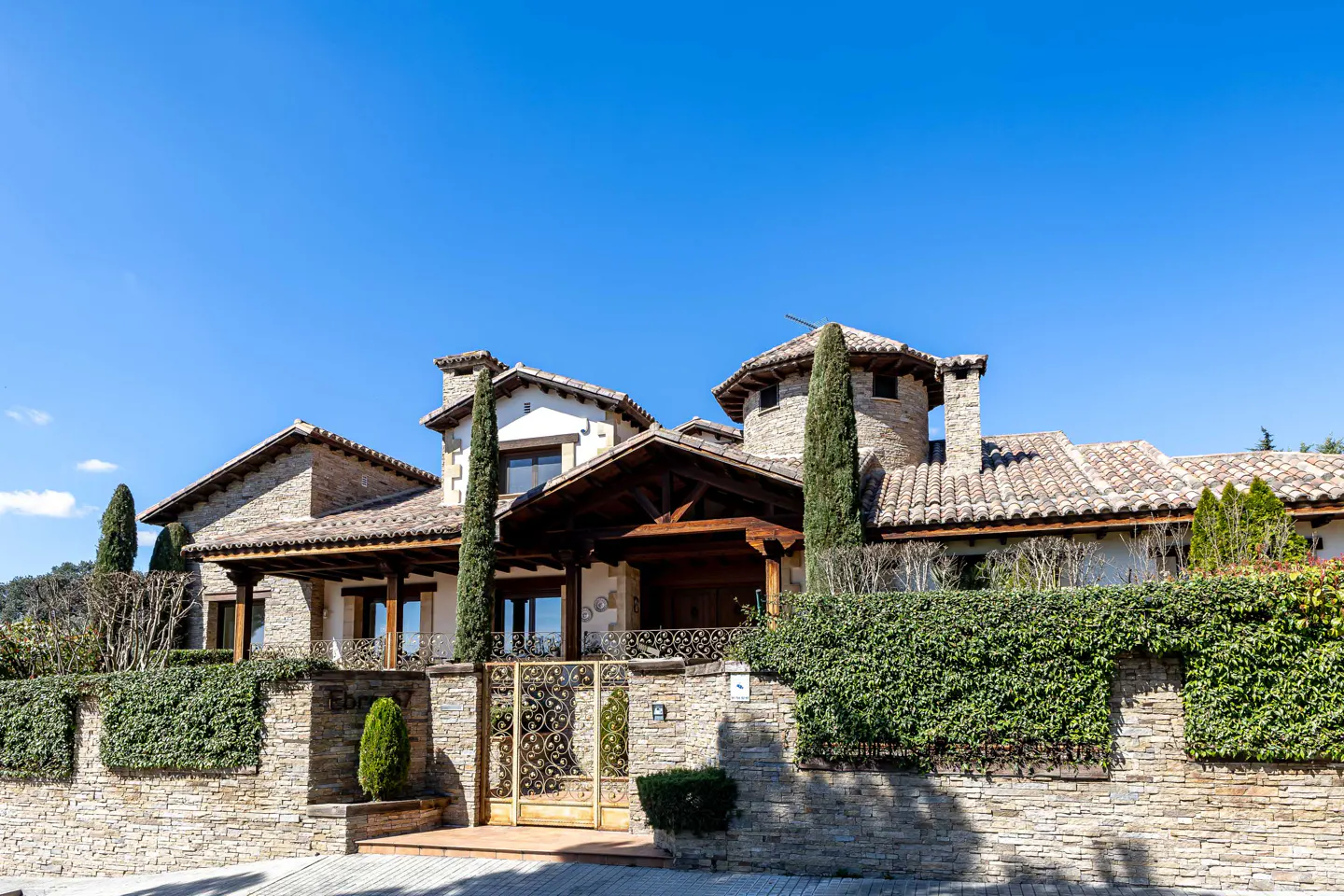 Exterior view of a stone house with a tile roof, a wooden porch, and a stone wall with a metal gate.