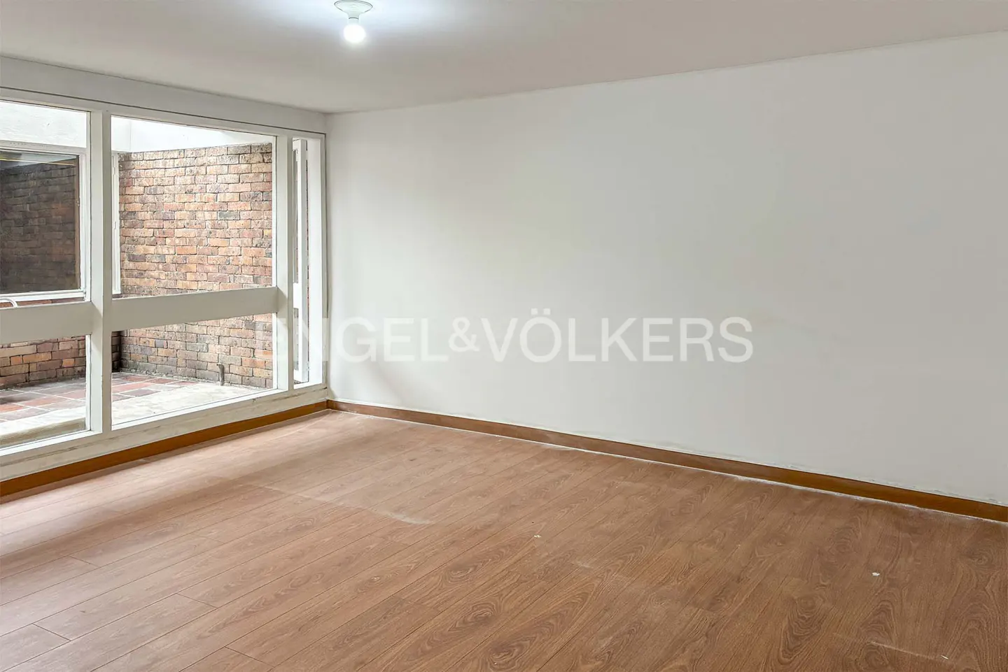 Empty room with wood floors, white walls, and large windows overlooking a brick patio. Engel & Völkers logo on the wall.