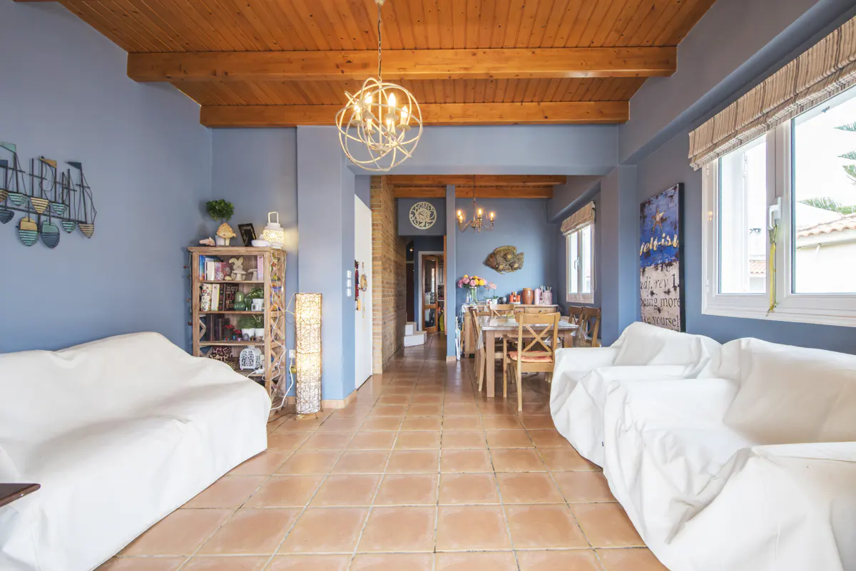 Living room with blue walls, wood ceiling, and terracotta tile floor. White sofas face each other. A dining table is visible in the background.