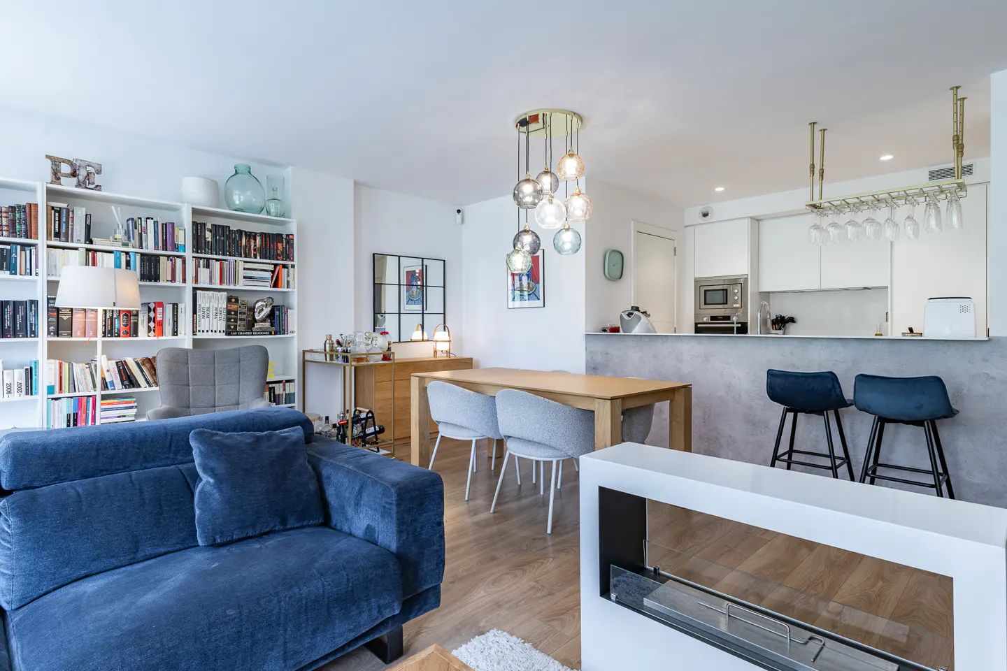 Open-plan living area with a blue sofa, dining table, modern pendant lights, and a kitchen with a breakfast bar.