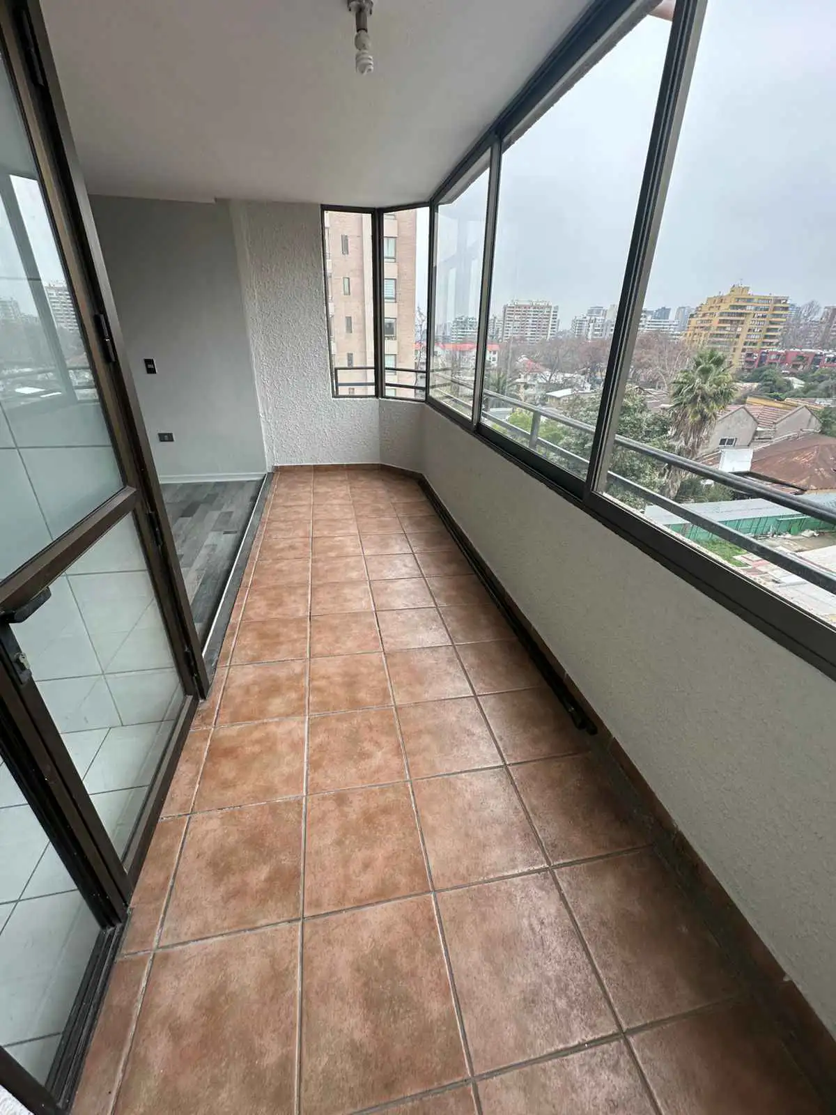 Empty balcony with brown tiled floor, large glass windows, and a view of the city skyline on a cloudy day.
