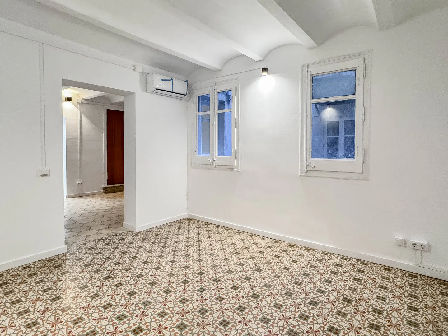 Bright, empty room with white walls, two windows, and patterned tile floor. An open doorway leads to another room.