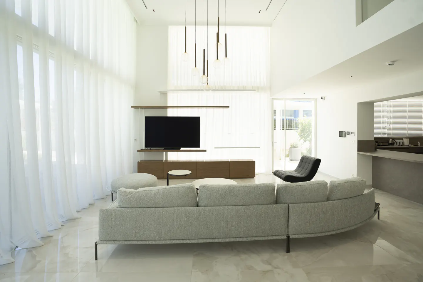 Bright, modern living room with a gray sofa, TV, and floor-to-ceiling white curtains. A black lounge chair sits near a sliding glass door.