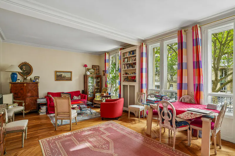 Bright living room with hardwood floors, red sofa, and dining table by large windows with colorful curtains.