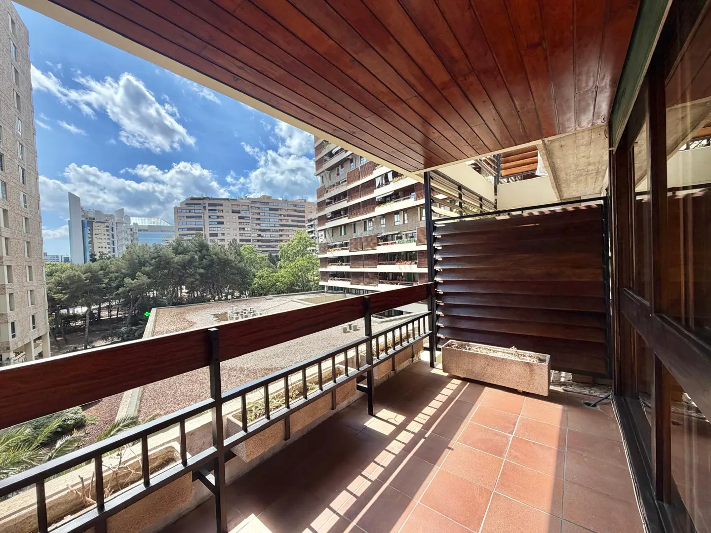Balcony view with brown wood ceiling, terracotta tile floor, and black metal railing. City buildings and blue sky visible in the background.
