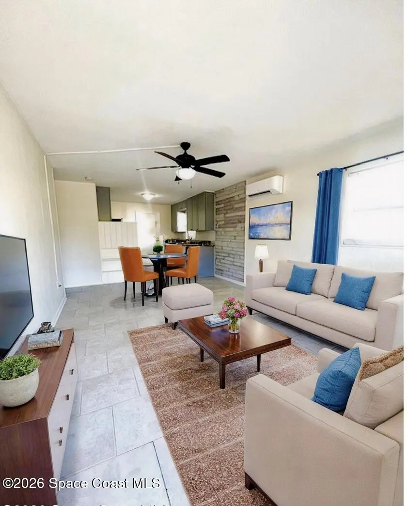 Open-concept living room with beige sofas, blue pillows, and a dark wood coffee table on a brown rug. Orange chairs surround a dining table.