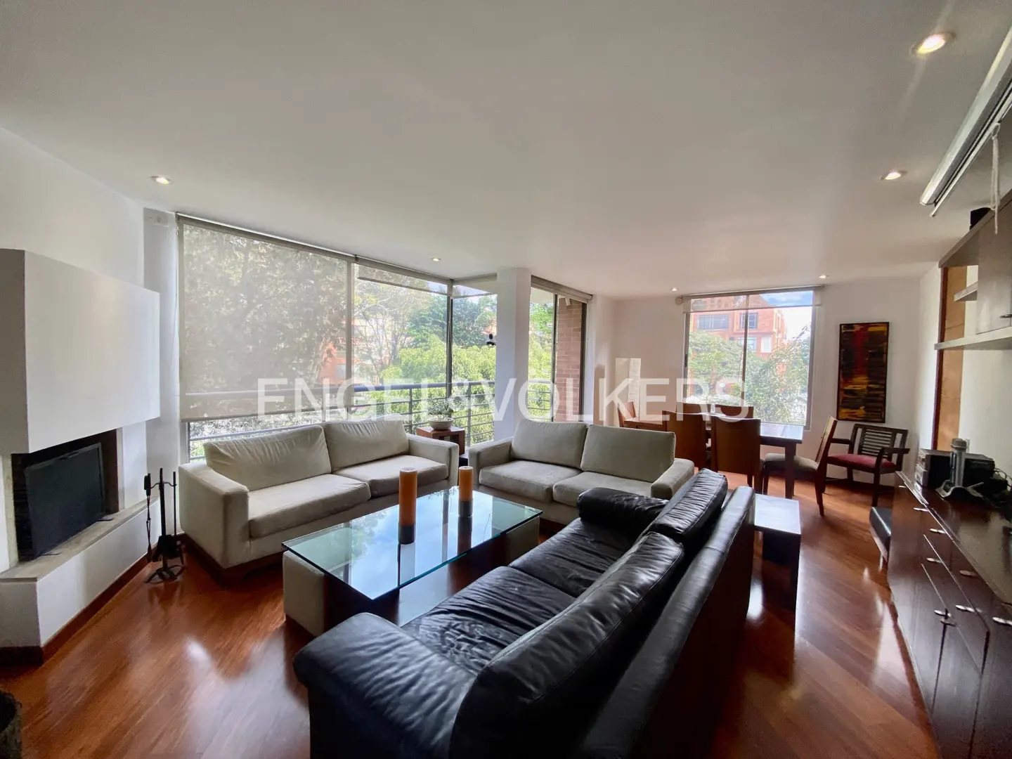 Bright living room with hardwood floors, white walls, and large windows overlooking greenery. Sofas, a glass table, and a fireplace add to the inviting space.