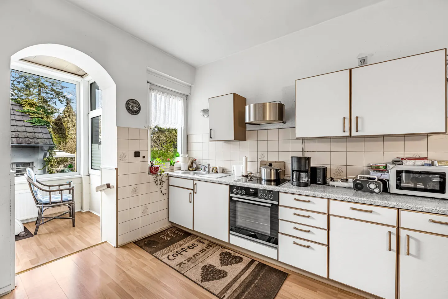 Bright kitchen with white cabinets, tiled backsplash, and wood floor. A chair sits in the sunroom visible through an arched doorway.