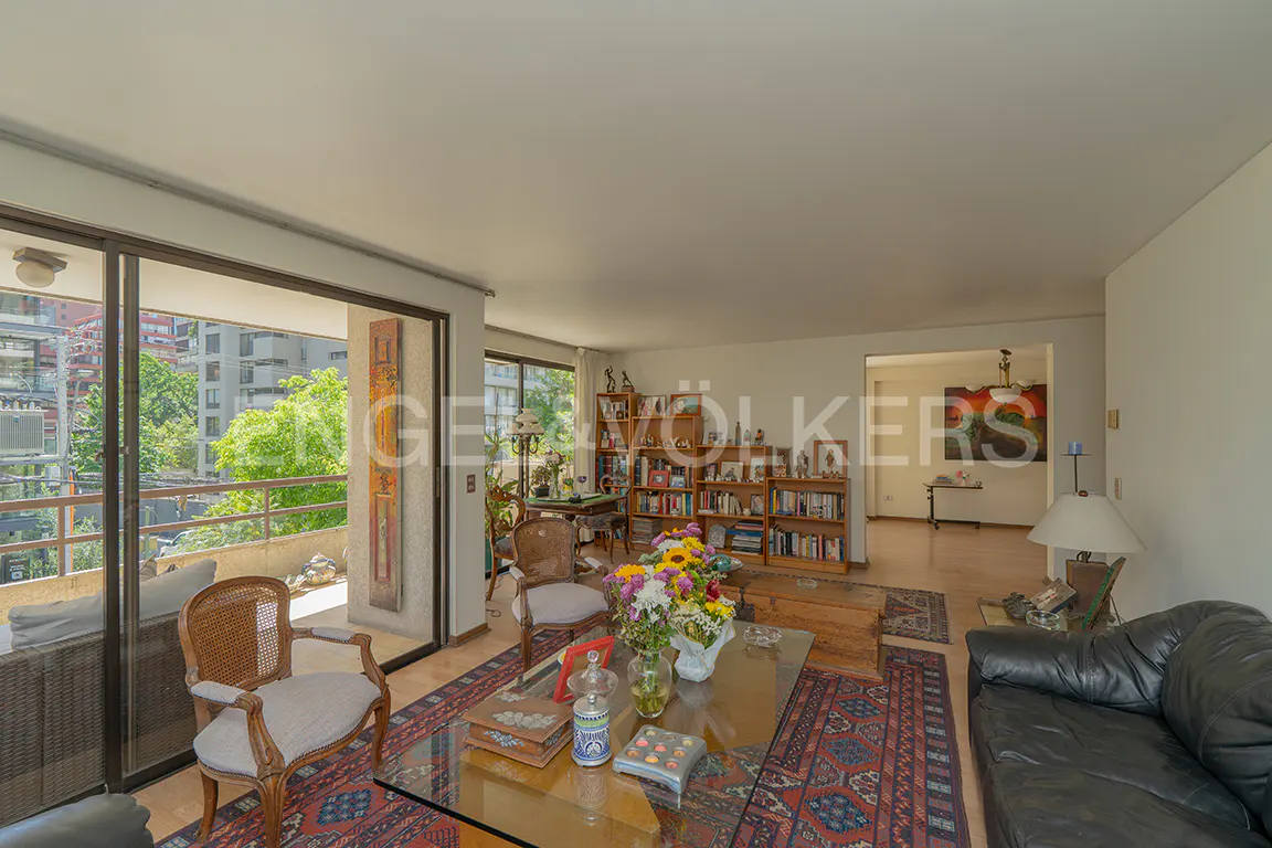 Bright living room with a glass coffee table, patterned rug, bookshelves, and a balcony with city views.