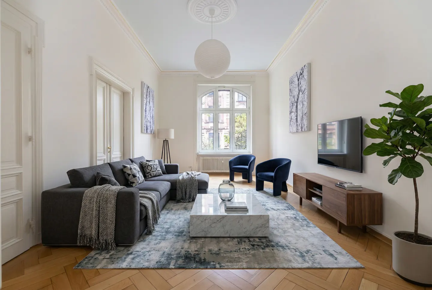 Bright living room with gray sectional, blue chairs, marble table, and wood floors. A large window provides natural light.
