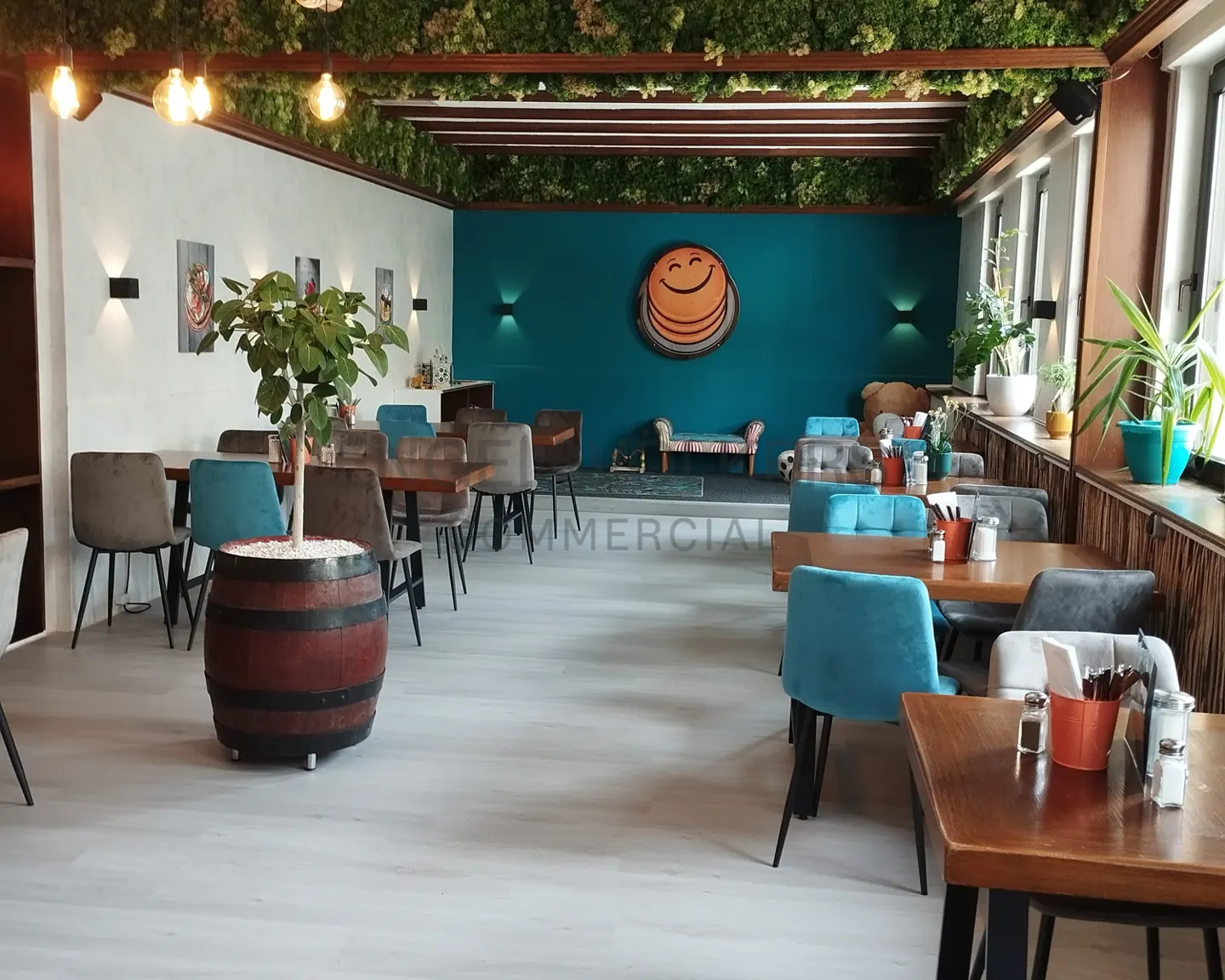 Restaurant interior with tables, chairs, and a barrel planter. Teal accent wall with a smiling pancake logo. Greenery hangs from the ceiling.