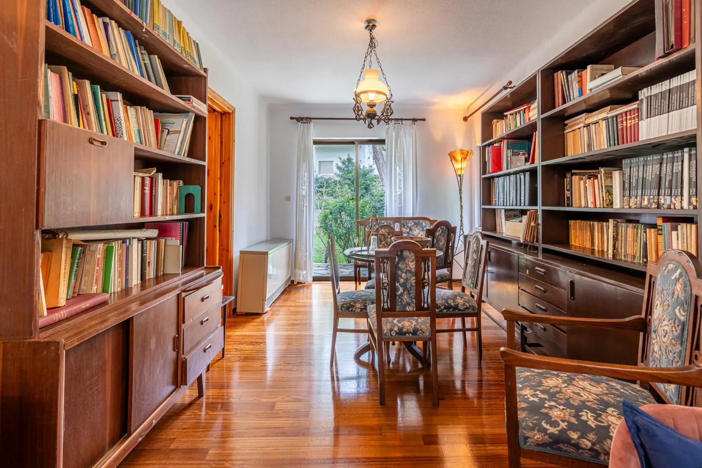A room with hardwood floors, bookshelves filled with books, a dining table with chairs, and a window with a view of greenery.