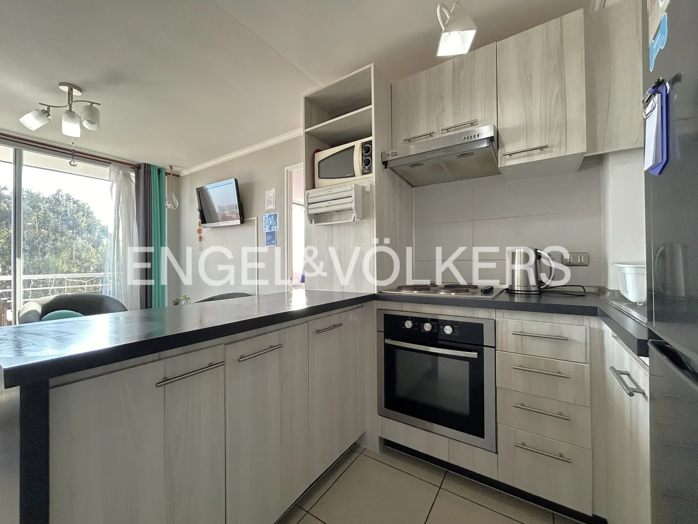 A modern kitchen with light wood cabinets, black countertops, and stainless steel appliances. A window overlooks trees.