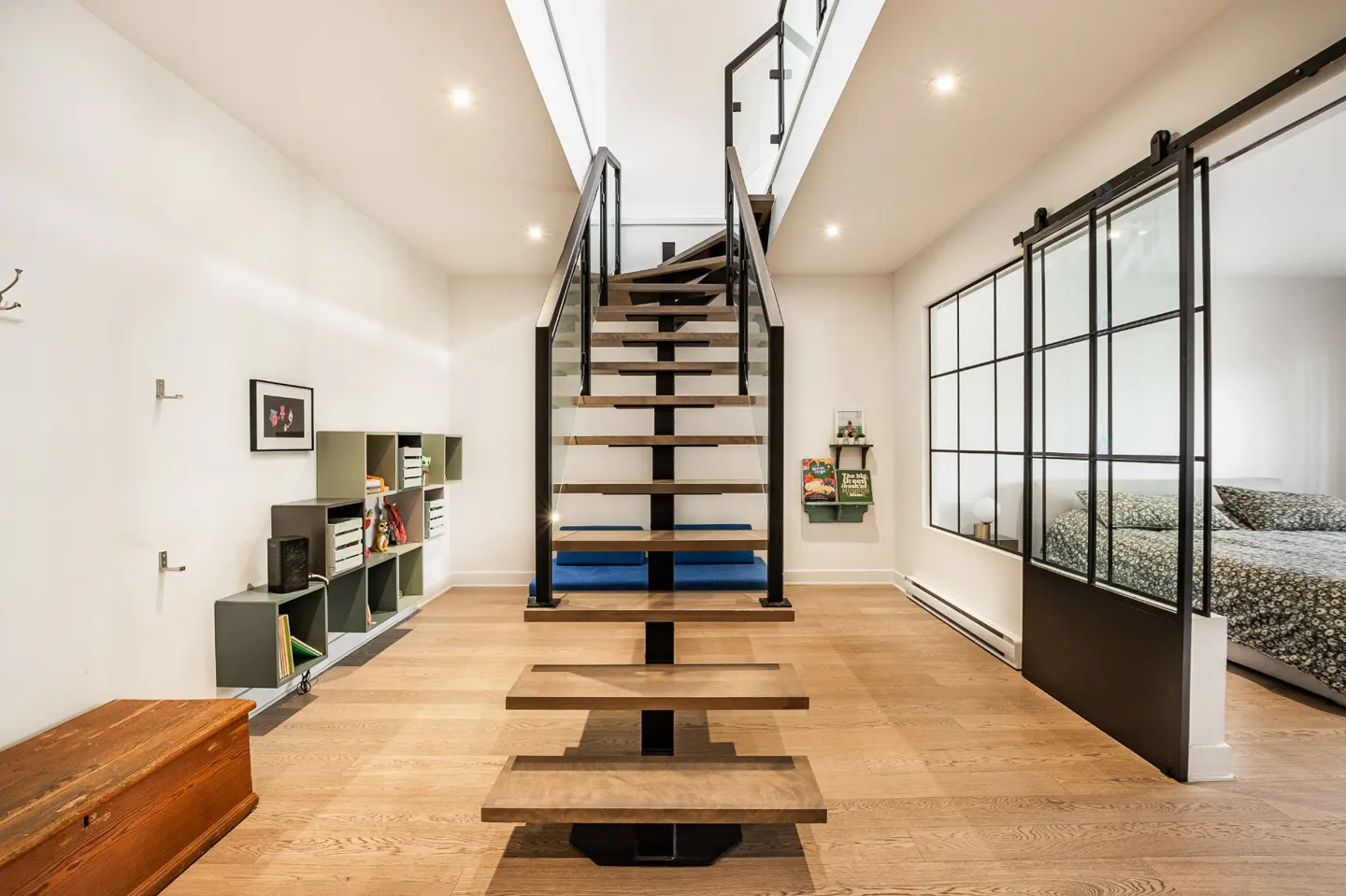 Modern interior with floating wood stairs, black metal railings, and glass panels. A sliding barn door with glass panes leads to a bedroom. Wood floors and white walls.