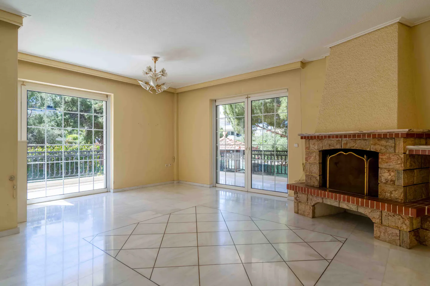 Bright, empty living room with marble floors, yellow walls, and a stone fireplace. Glass doors lead to a balcony with greenery visible outside.