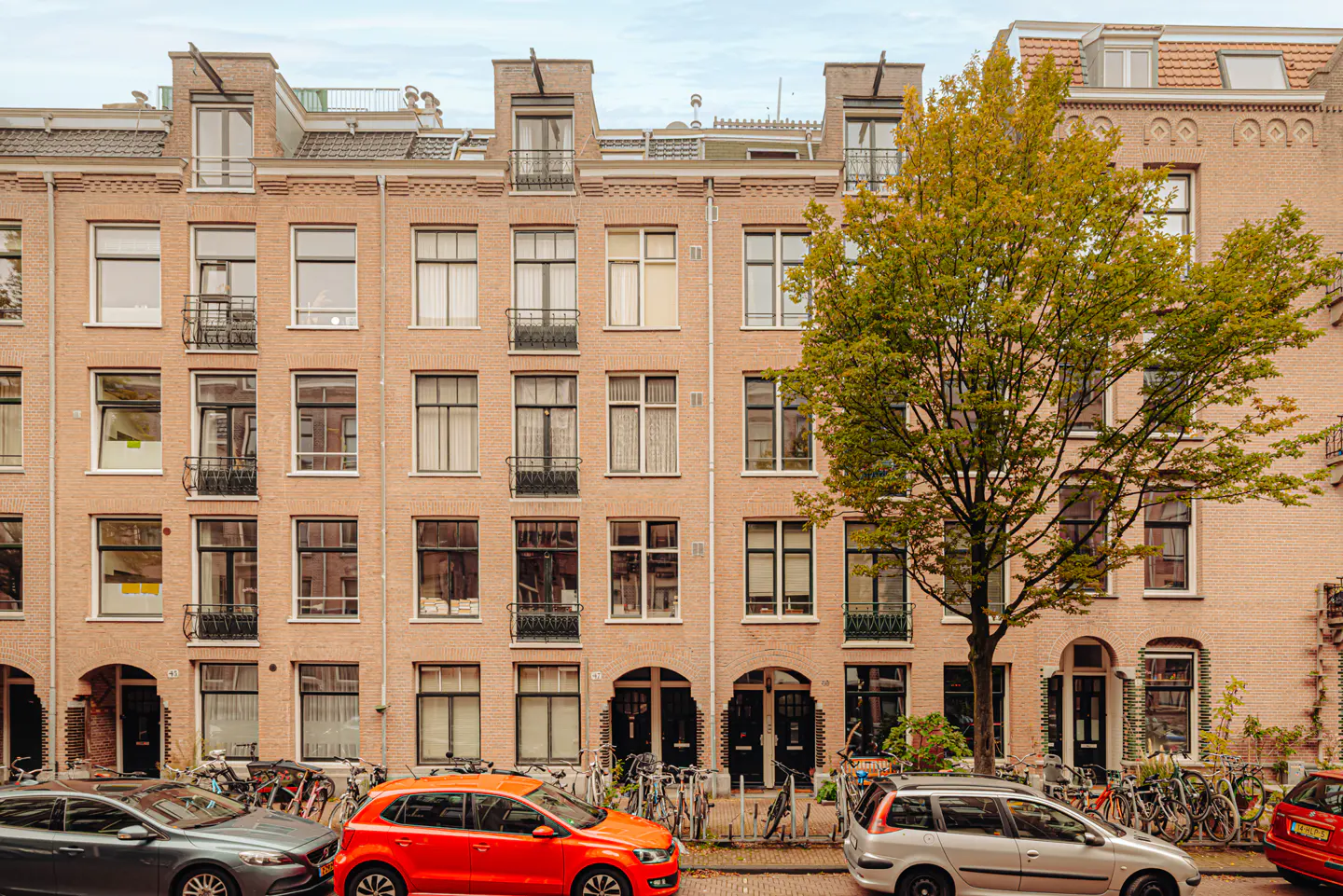 Row of brick townhouses with dark windows and doors, parked cars and bikes line the street. A tree with green leaves is on the right.