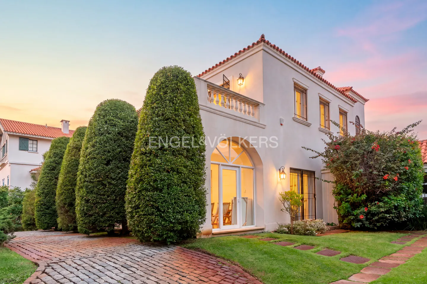 Exterior of a two-story white house with a red tile roof, green bushes, and a brick driveway at sunset.