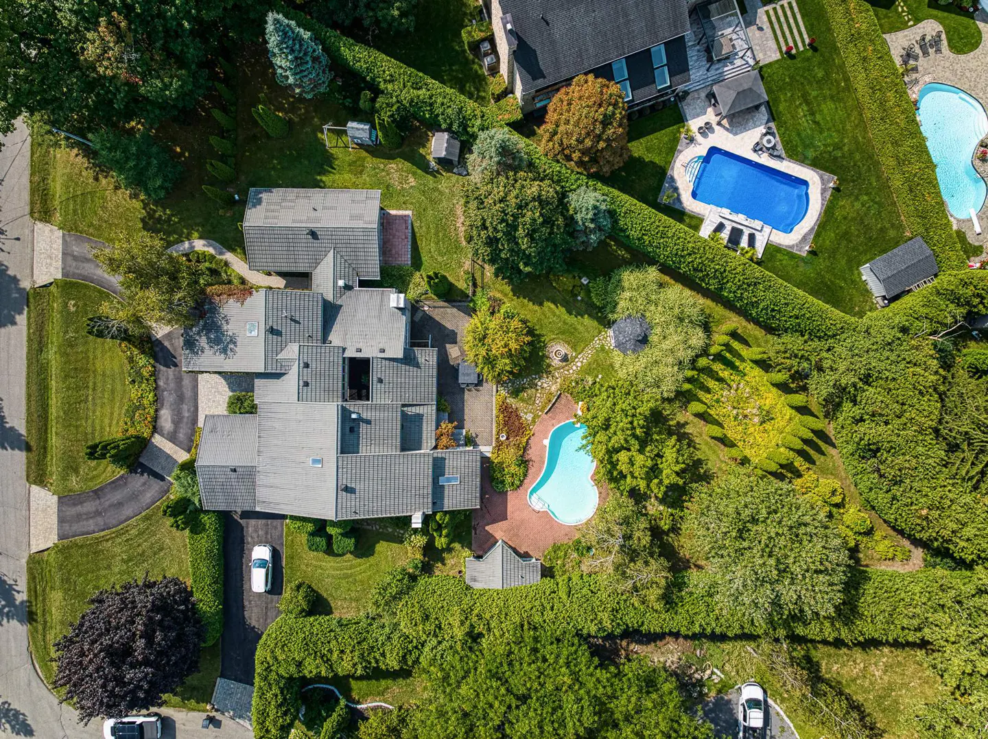 Aerial view of a large house with a gray roof, surrounded by green trees and hedges, with two blue swimming pools.