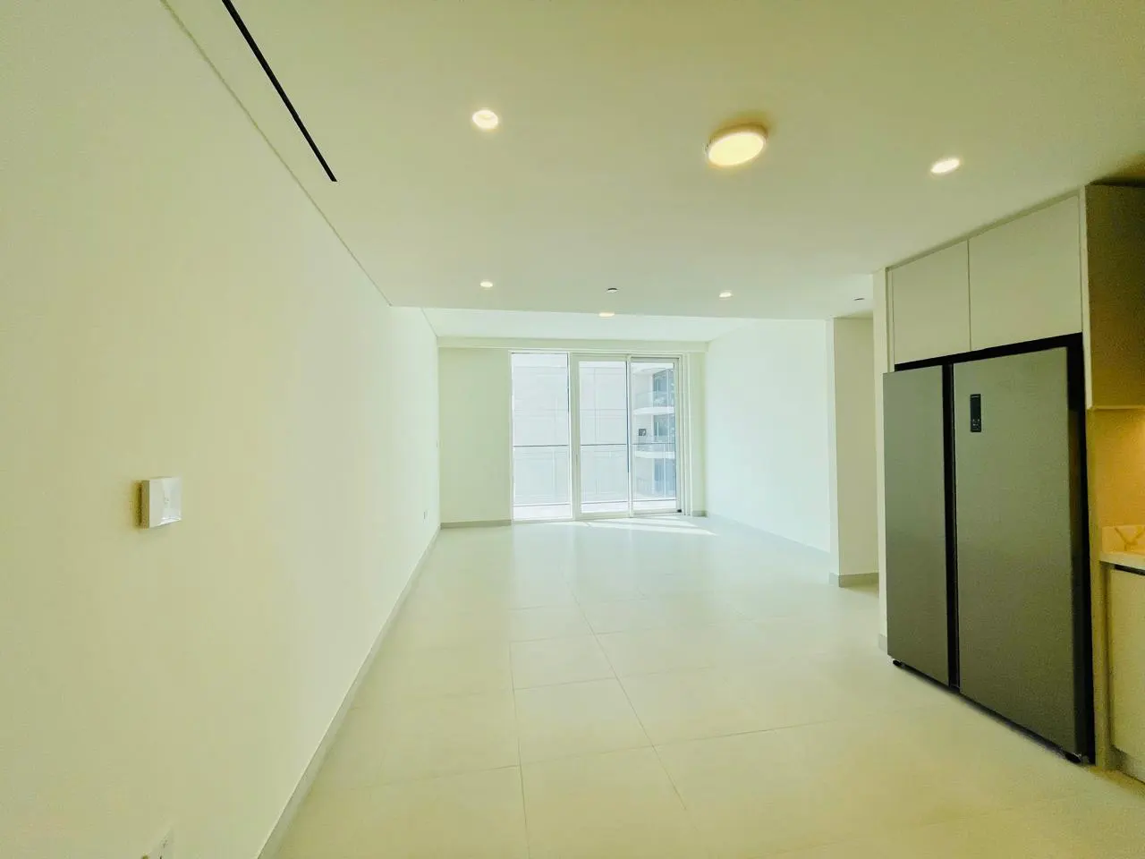 Bright, empty apartment with white walls and tile floors. Sliding glass doors lead to a balcony. A stainless steel refrigerator stands near white cabinets.