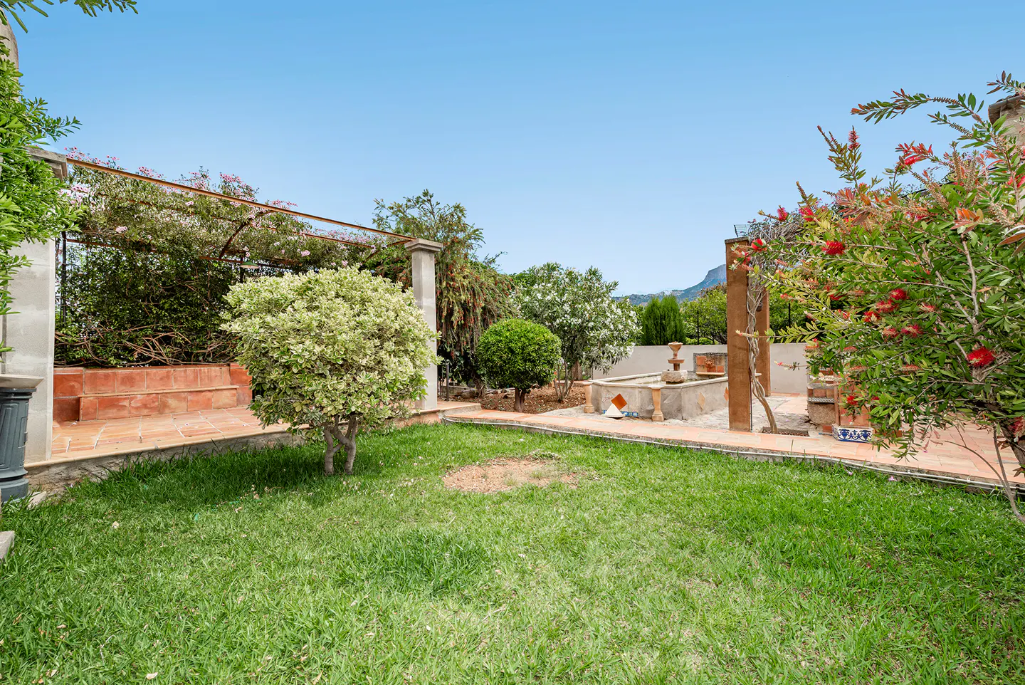 A lush green lawn with trees, flowers, and a stone fountain under a clear blue sky.