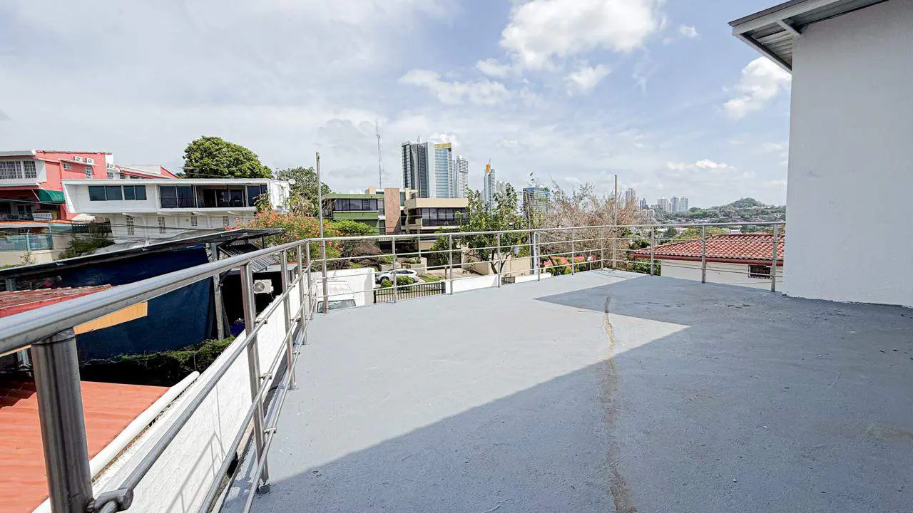 Wide shot of a gray rooftop patio with a metal railing, overlooking a cityscape with buildings and trees under a cloudy sky.