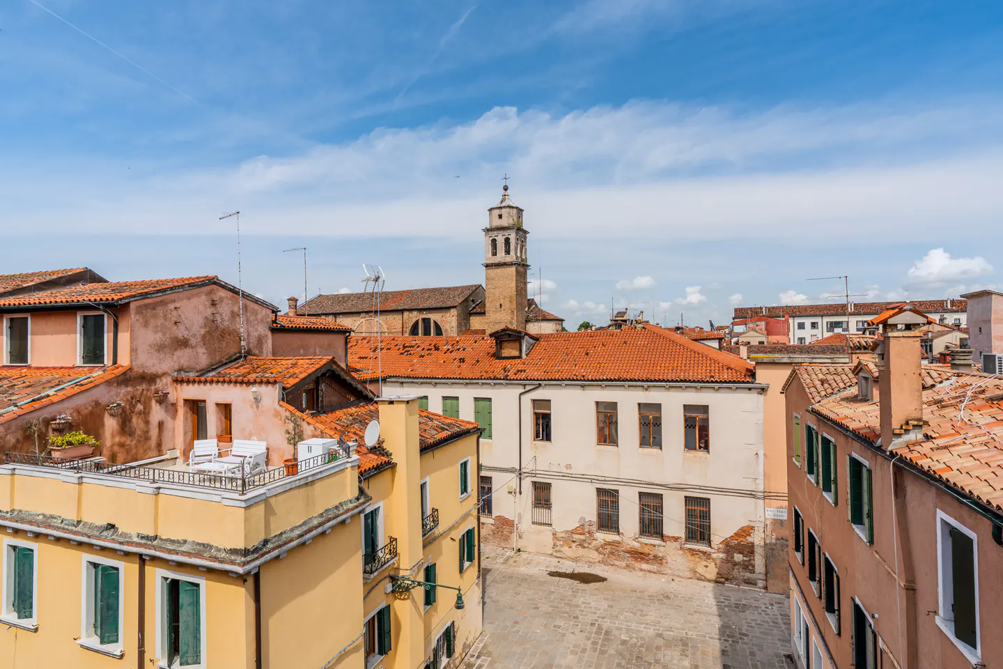 Venice cityscape with terracotta rooftops, yellow buildings, and a church tower under a blue sky.
