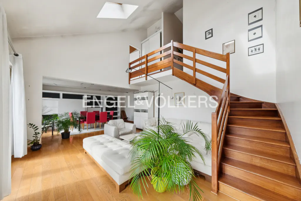 Bright, modern living room with wooden floors, white walls, and a wooden staircase. White sofa and chairs, with a red dining set in the background.