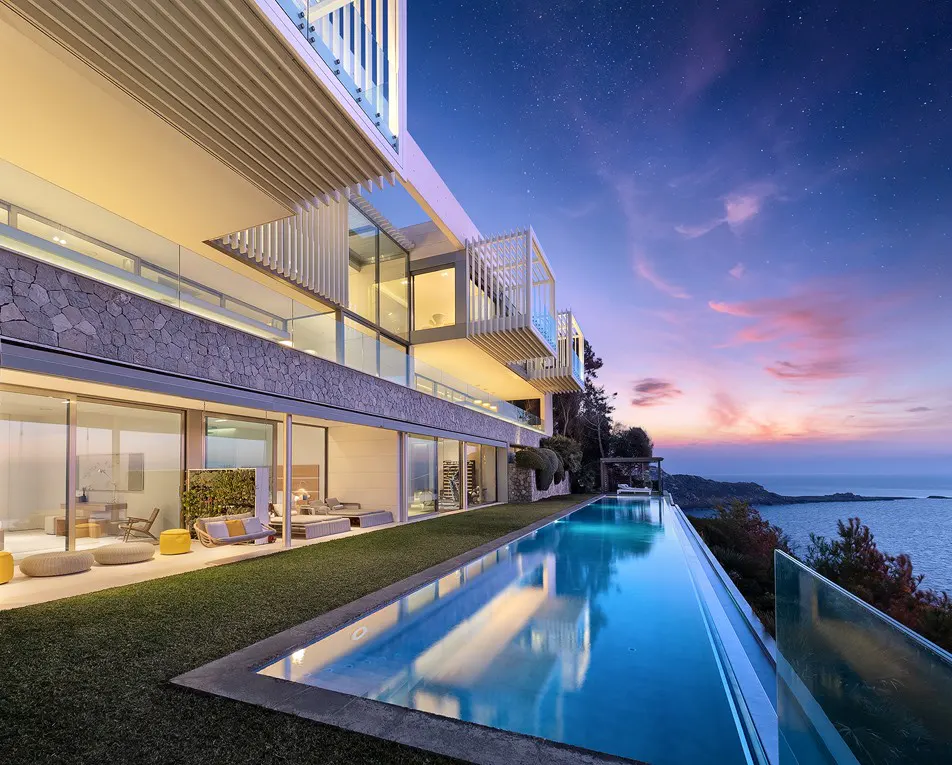 Modern luxury home with an infinity pool overlooking the ocean at dusk. The sky is a mix of blue, purple, and pink.