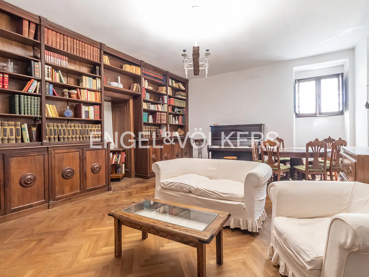 A living room with a large wooden bookcase filled with books, a white sofa, a wooden coffee table, a piano, and a dining table.