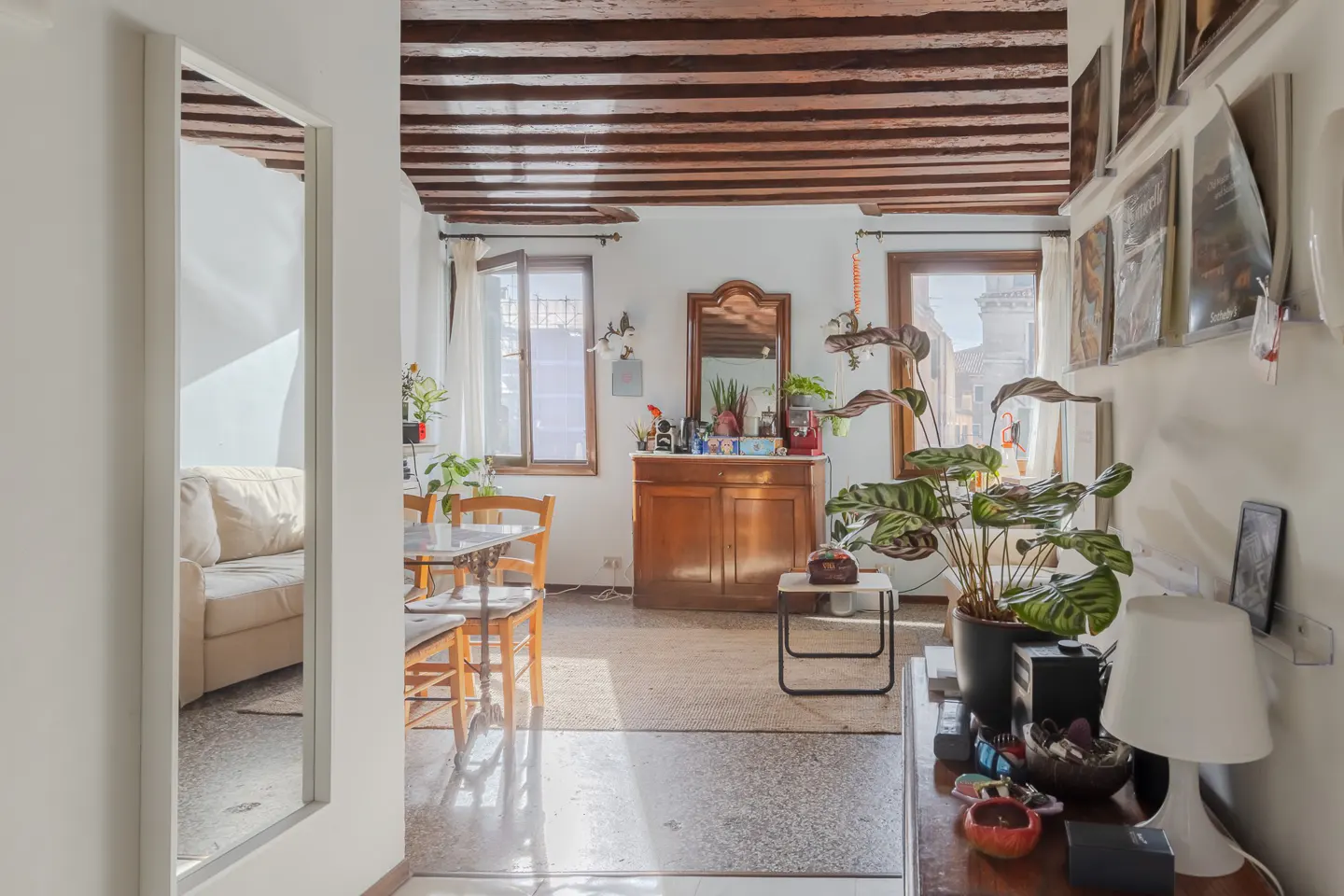 A bright living room with exposed wood beams, white walls, and a dining table. A mirror reflects a cream sofa. Plants add a touch of nature.