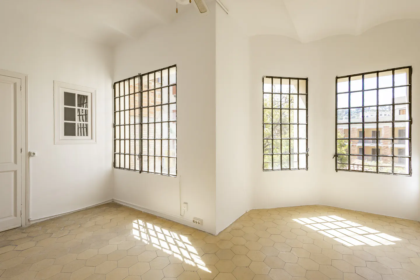 Bright, empty room with white walls, hexagonal tile floor, and barred windows letting in sunlight. A white door is on the left.