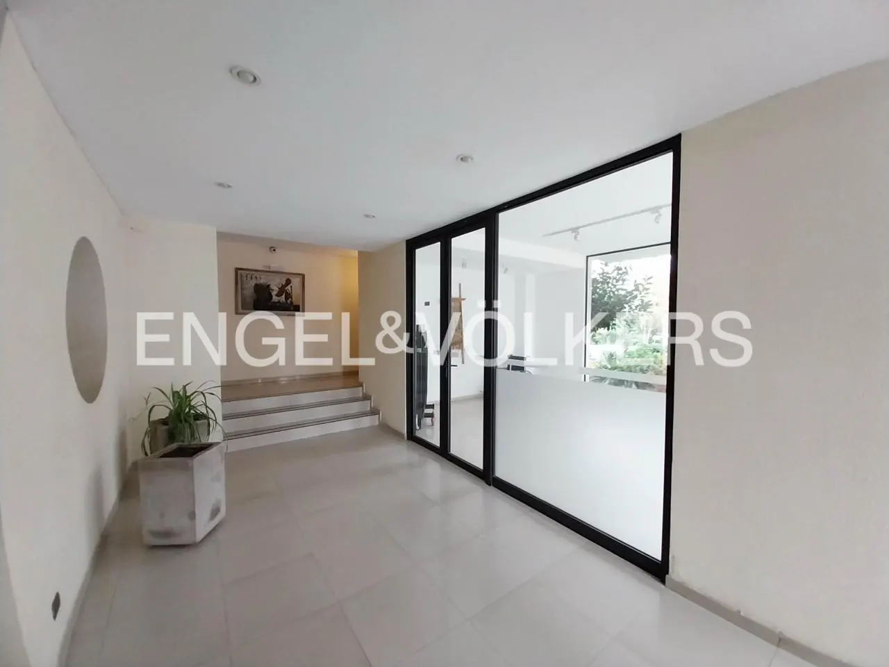 A bright, modern real estate office with tile floors, a plant, and a glass-walled office. The Engel & Volkers logo is visible.