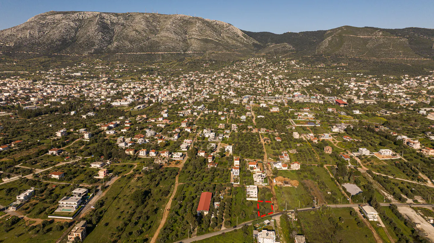 Aerial view of a property lot outlined in red, nestled in a green, hilly landscape with a town and mountain backdrop.