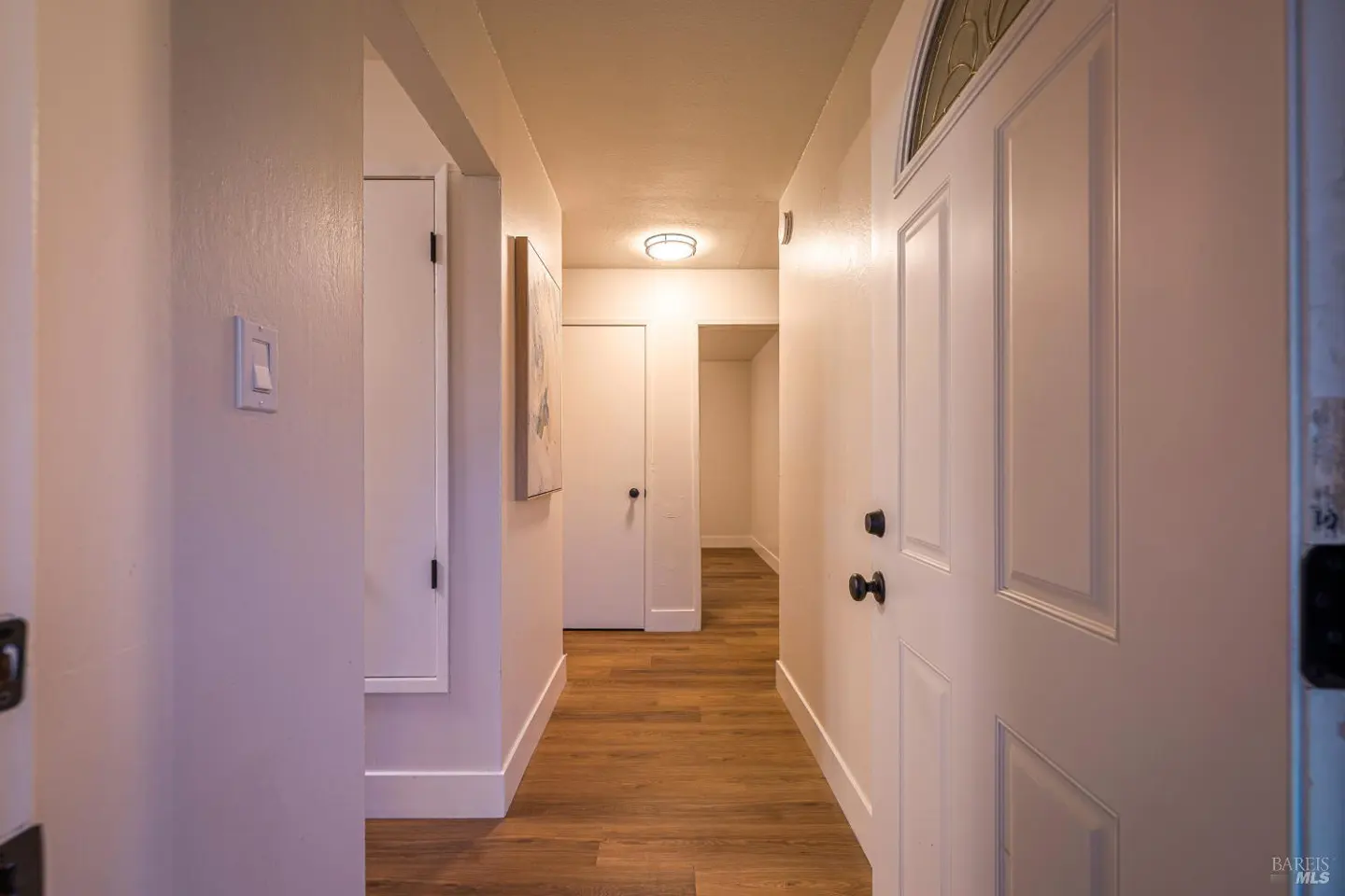 Interior shot of a hallway with wood floors, white walls, and white doors. A light fixture is visible on the ceiling.