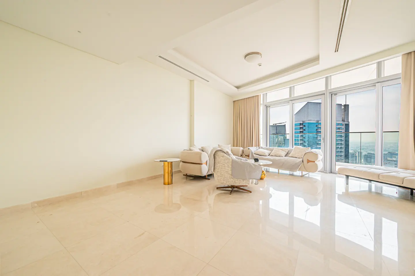 Bright, modern living room with beige sofas, a swivel chair, and floor-to-ceiling windows overlooking a city skyline.