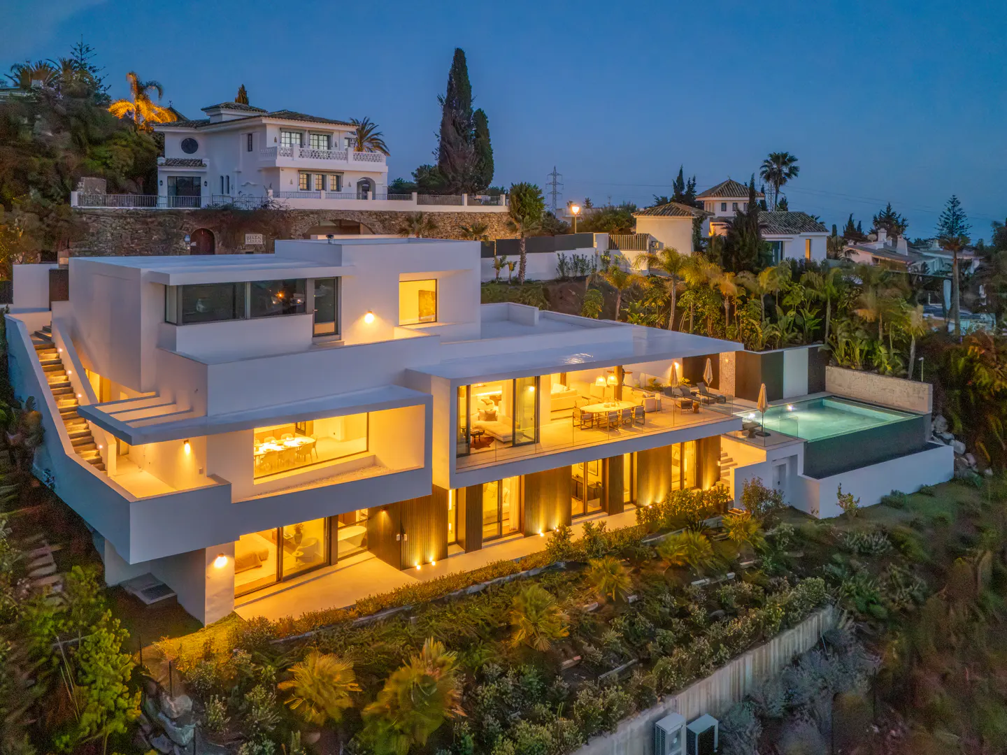 Modern white villa at dusk, with large windows glowing with warm light, a pool, and lush greenery.