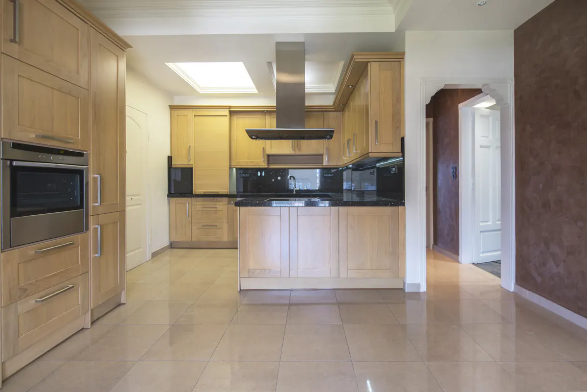 A bright kitchen with light wood cabinets, black countertops, and stainless steel appliances. A doorway leads to another room with brown textured walls.
